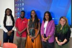 5 A group of five people standing in a room, smiling in front of a large screen displaying a computer desktop. They are dressed in various professional attire.