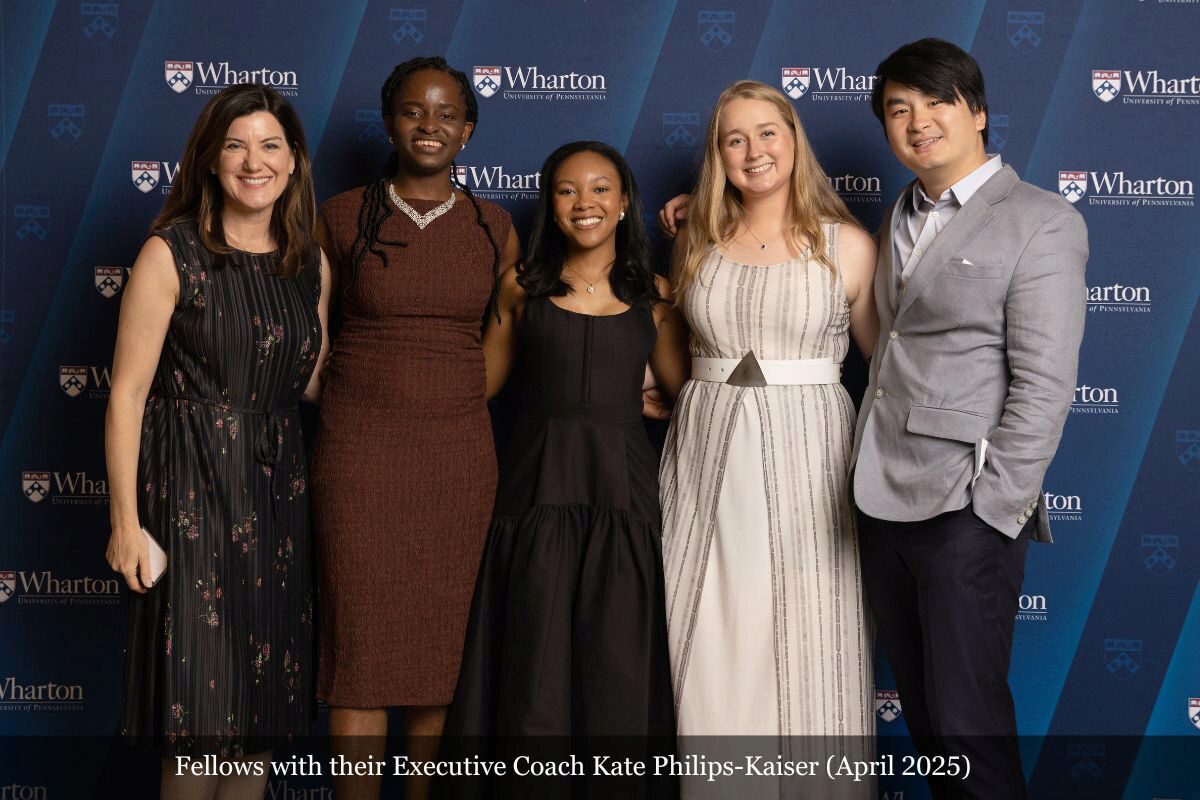A small group of people, including four individuals and their executive coach, is standing together, smiling in front of a Wharton School backdrop. They are dressed in formal attire.