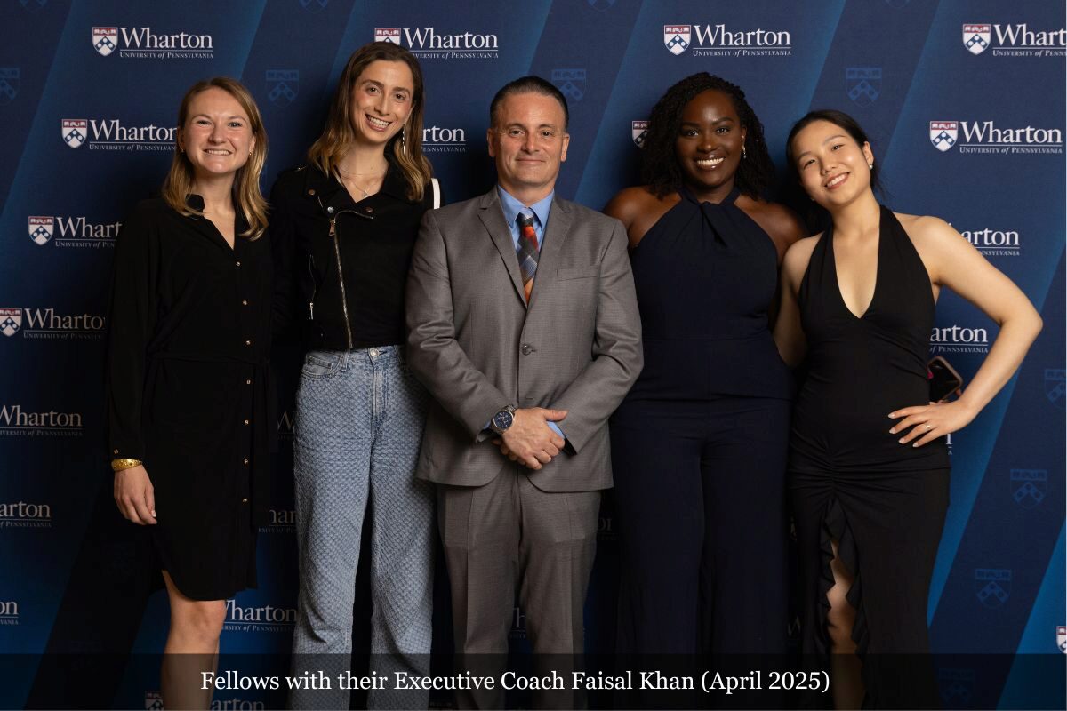 A group photo of several people standing in front of a Wharton School backdrop. They are dressed in formal and semi-formal attire, smiling at the camera.