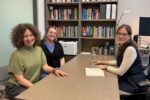 Three people are sitting and smiling in an office setting with bookshelves filled with books in the background. They appear to be having a casual meeting.
