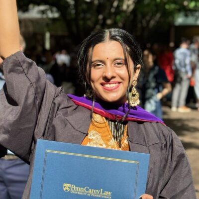 Lakshana Ramakrishnan Profile A graduate wearing academic regalia holds a diploma from Penn Carey Law, University of Pennsylvania, smiling in an outdoor setting.