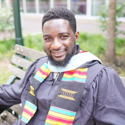 Lusayo Graduation Profile A person in a graduation gown and colorful stole, seated on a bench, smiling, representing academic achievement and celebration.